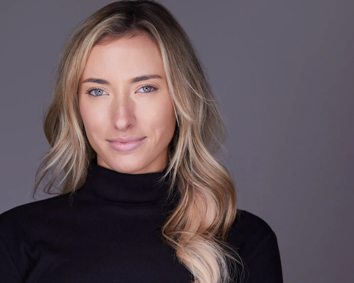 Actor headshot of a woman with long wavy blonde hair in a black turtleneck against a gray backdrop