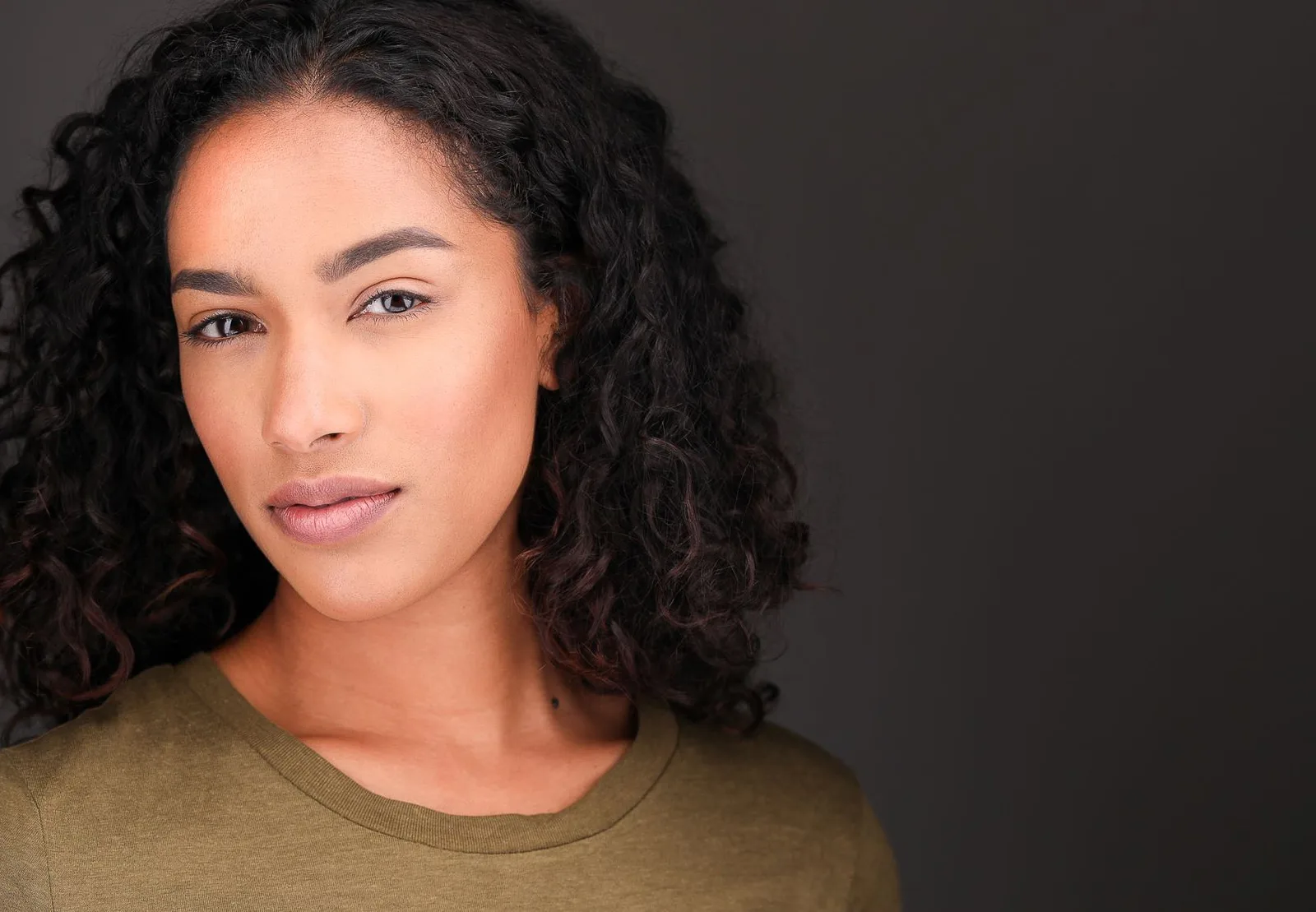 Actor headshot of a woman with long dark curly hair in an olive T-shirt against a charcoal backdrop