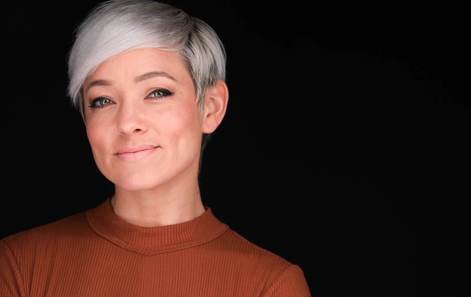 Actor headshot of a smiling woman with a silver pixie cut in a rust ribbed sweater against a black backdrop