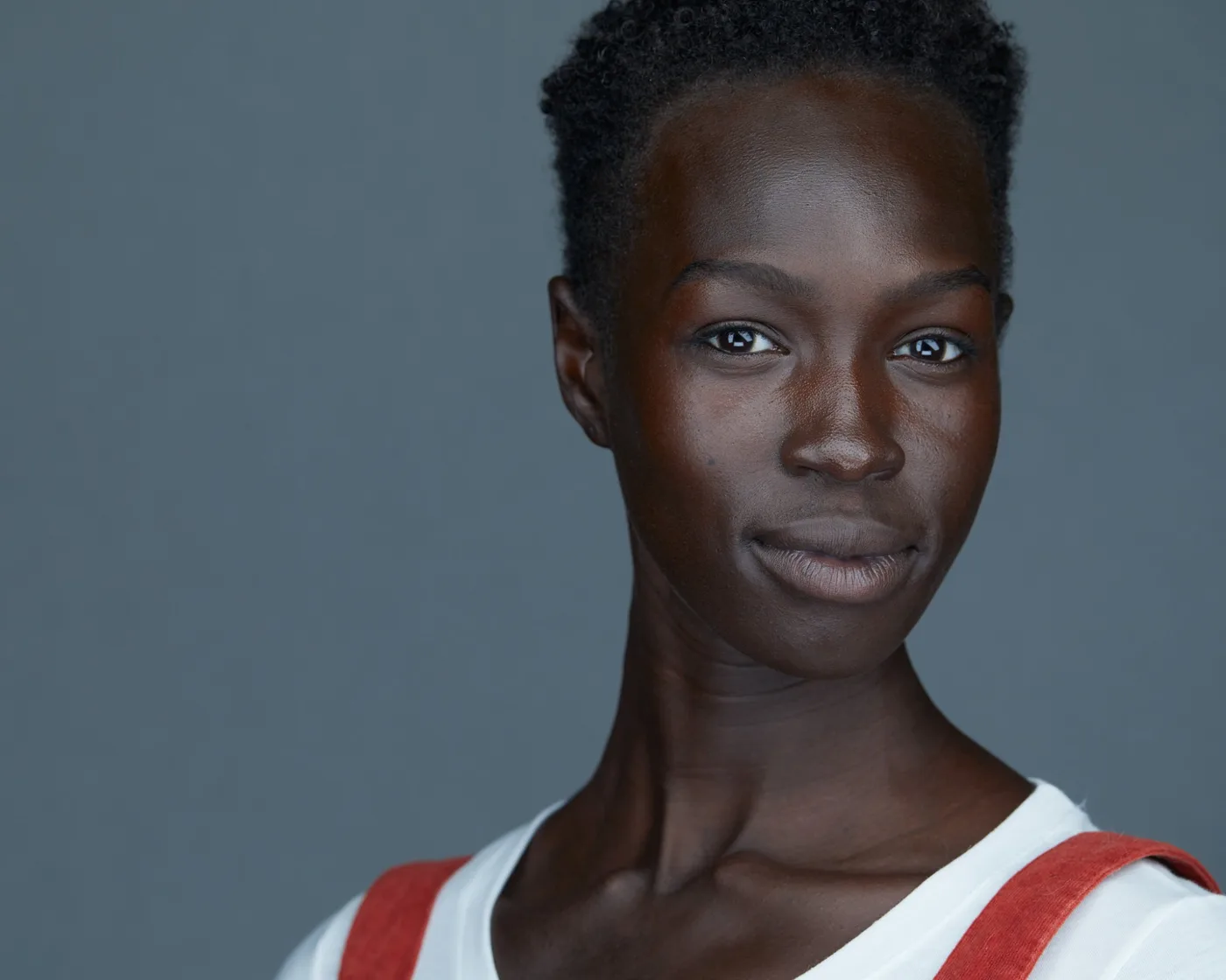 Actor headshot of a woman with short natural hair in a white T-shirt and rust overall straps against a slate backdrop