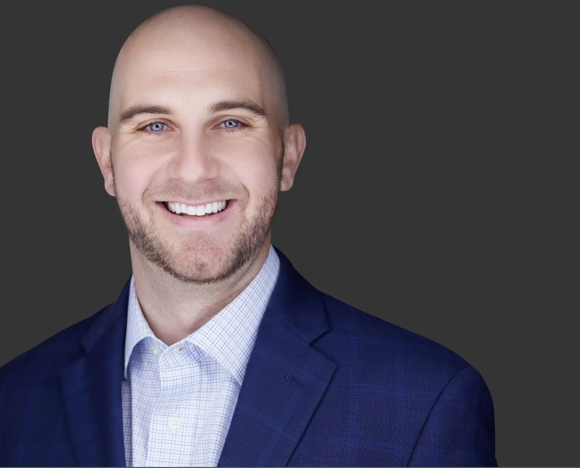 Business headshot of a smiling man with a shaved head and beard in a navy suit
