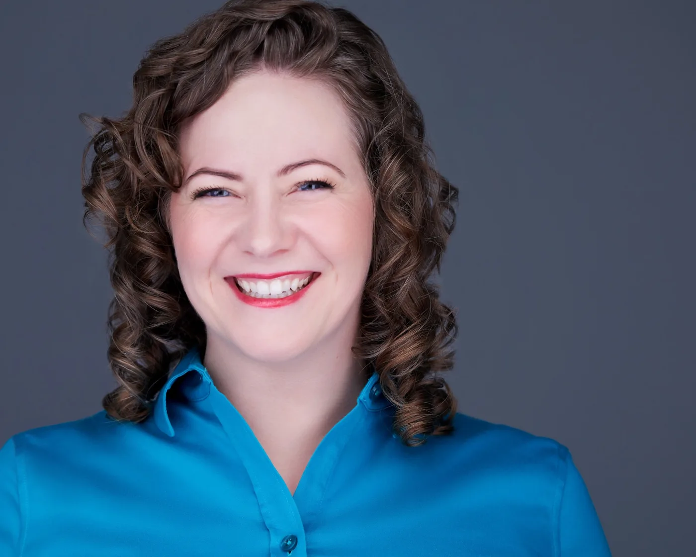 Business headshot of a smiling woman with curly brown hair in a teal blouse against a slate backdrop