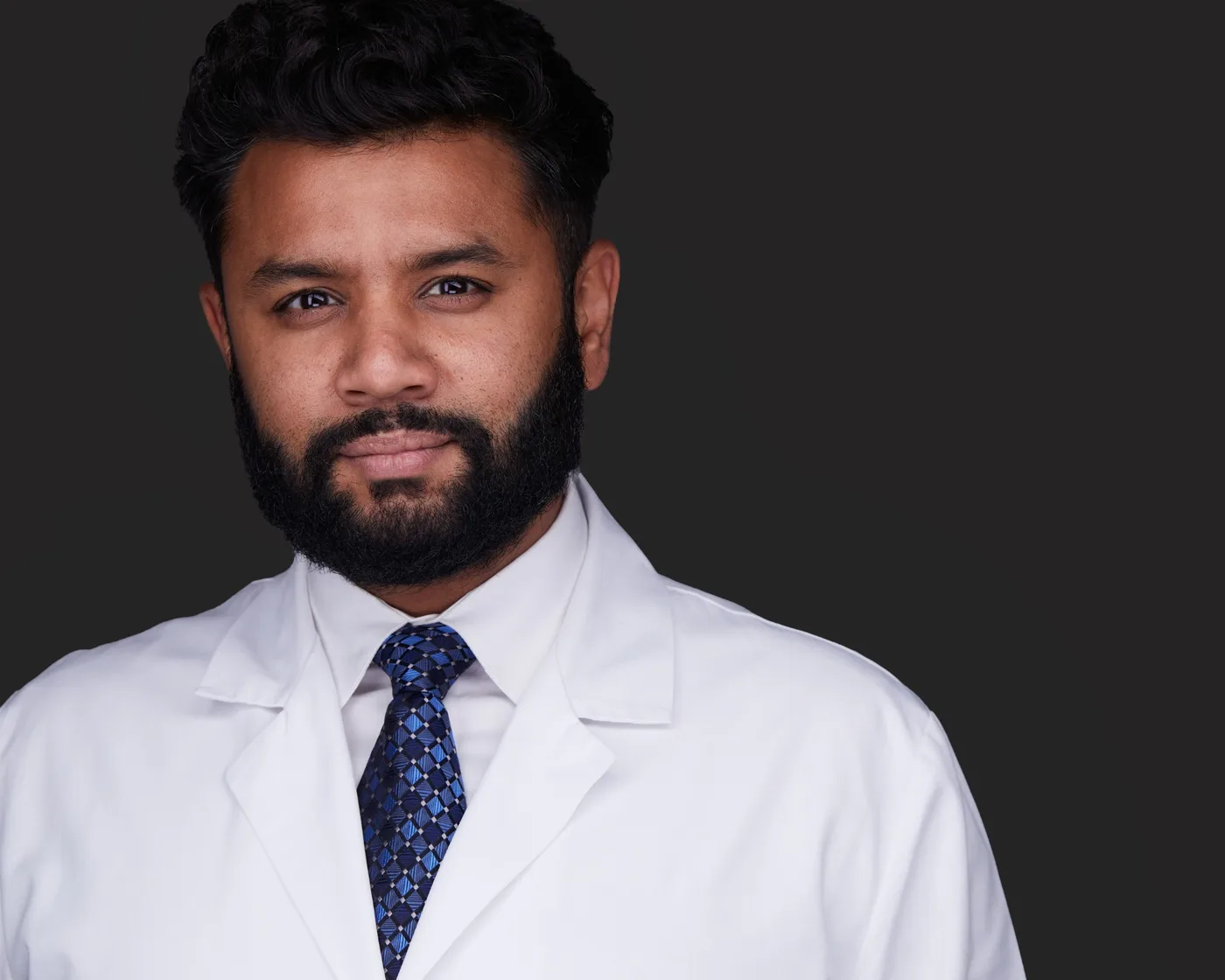 Business headshot of a man with a full beard in a white coat and blue patterned tie against a charcoal backdrop