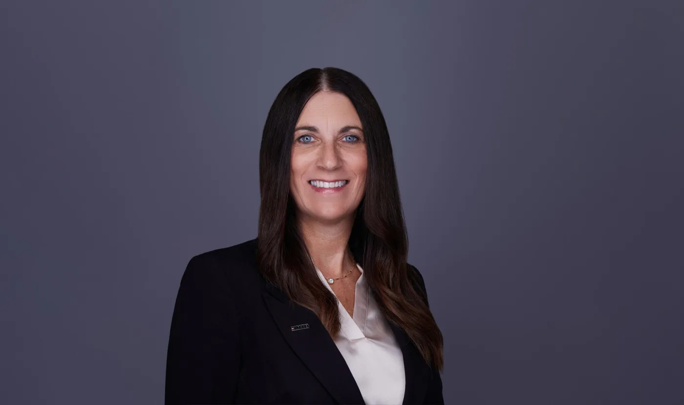 Corporate team headshot of a woman with long dark hair in a black blazer and ivory blouse against a gray backdrop