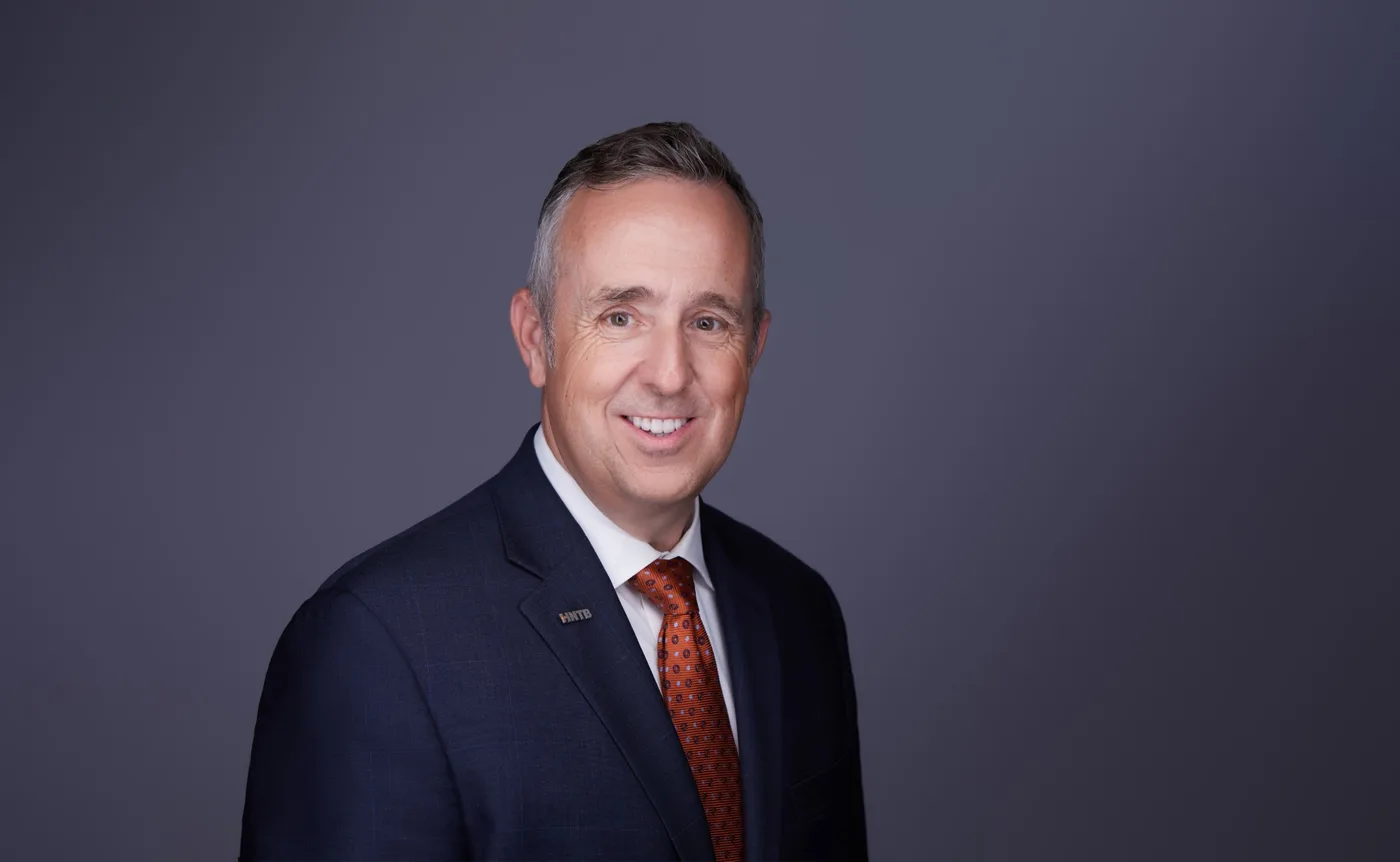 Corporate team headshot of a man with short graying hair in a navy suit and rust patterned tie against a gray backdrop