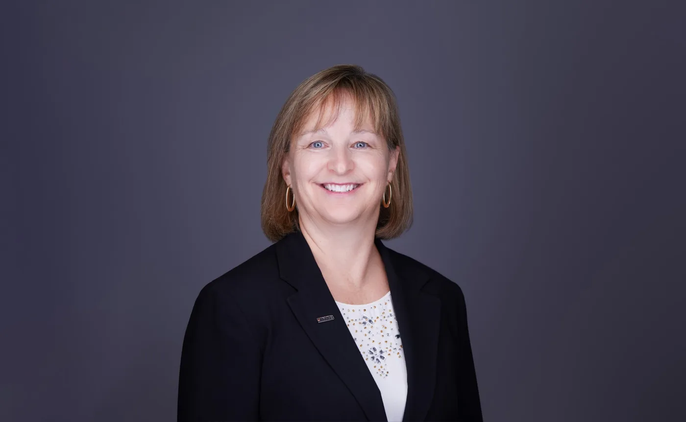 Corporate team headshot of a woman with a shoulder-length bob and hoop earrings in a black blazer against a gray backdrop