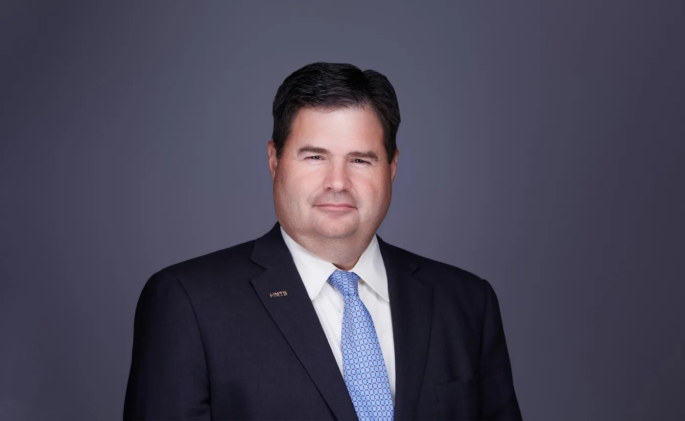 Corporate team headshot of a man with dark hair in a charcoal suit and light blue tie against a gray backdrop
