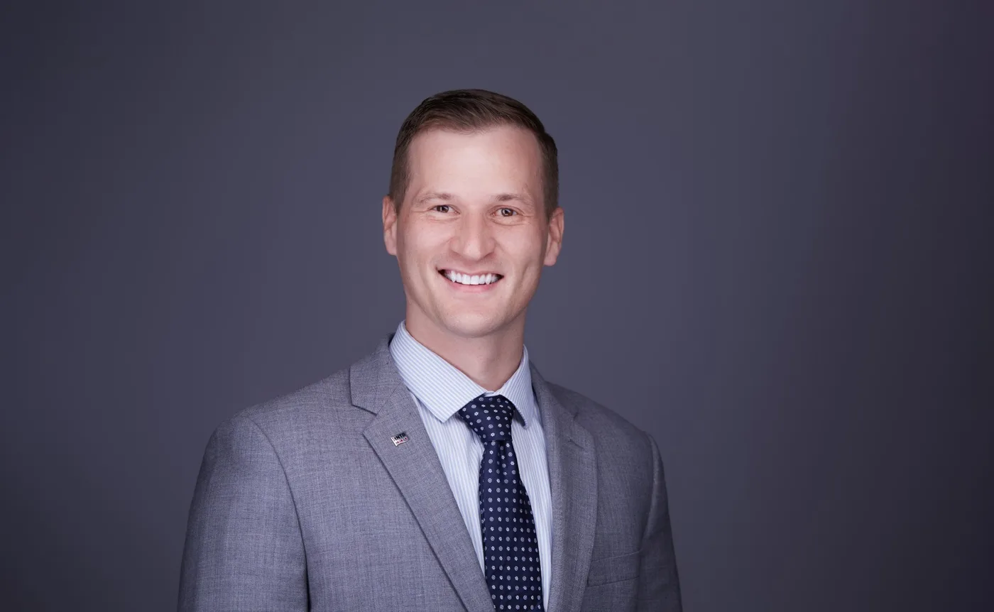 Corporate team headshot of a man with short brown hair in a light gray suit and navy dotted tie against a gray backdrop