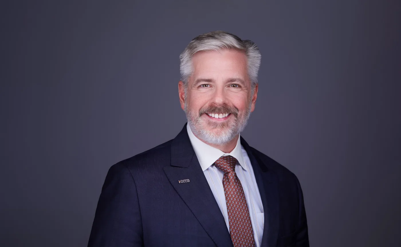 Corporate team headshot of a man with silver hair and a beard in a navy suit and burgundy tie against a gray backdrop