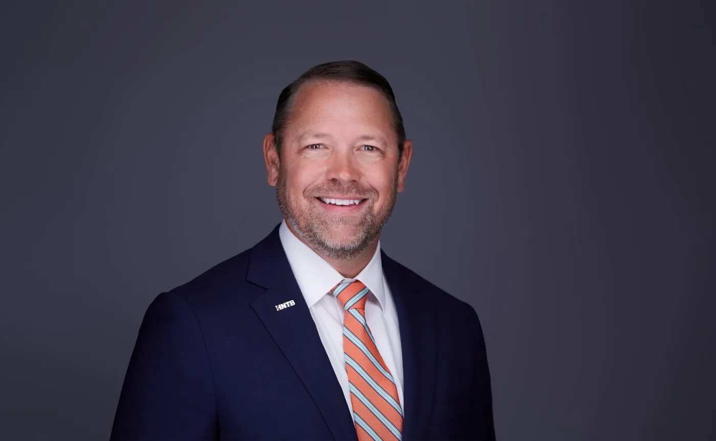 Corporate team headshot of a man with a short beard in a navy suit and orange striped tie against a gray backdrop