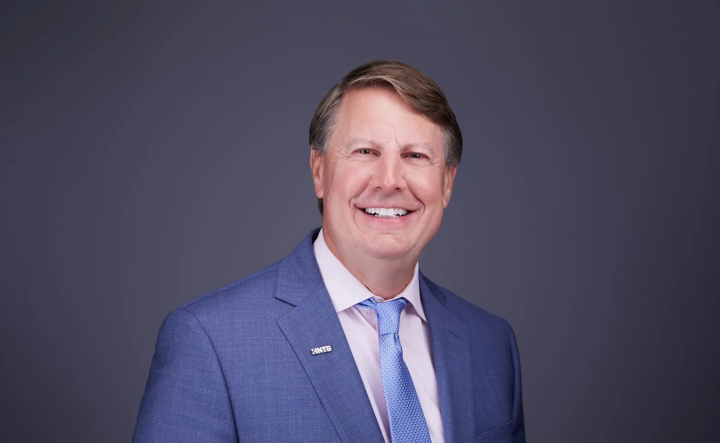 Corporate team headshot of a man with swept-back hair in a blue suit and light blue tie against a gray backdrop