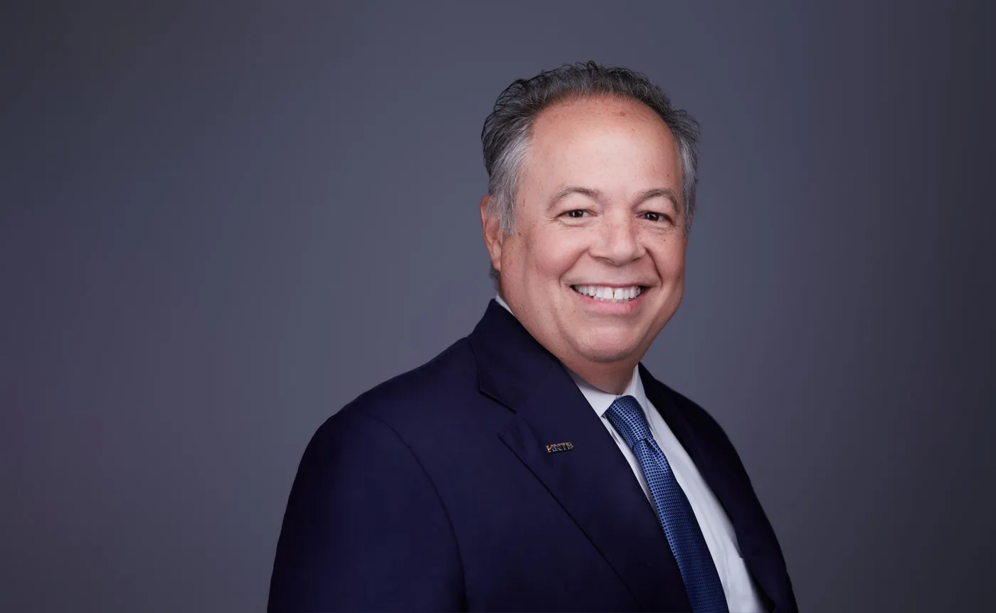 Corporate team headshot of a man with graying hair in a navy suit and blue tie against a gray backdrop