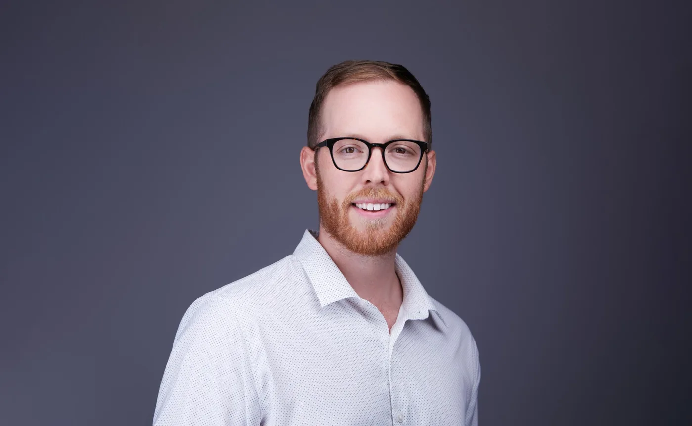 Corporate team headshot of a man with glasses and a red beard in a white patterned shirt against a gray backdrop